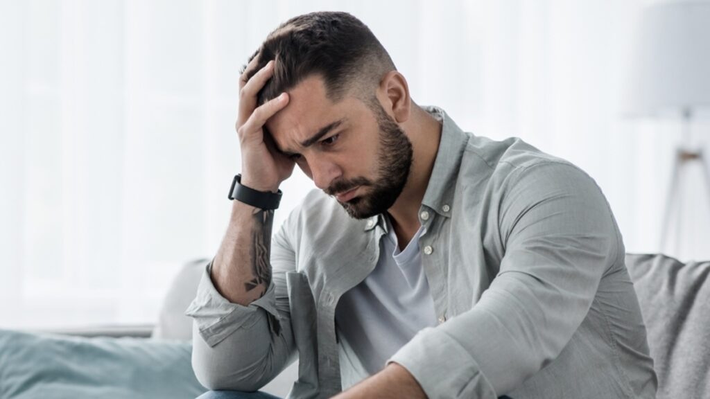 Bipolar Disorder in Adults: Adult man with bipolar disorder sitting on couch with head in hand looking distressed