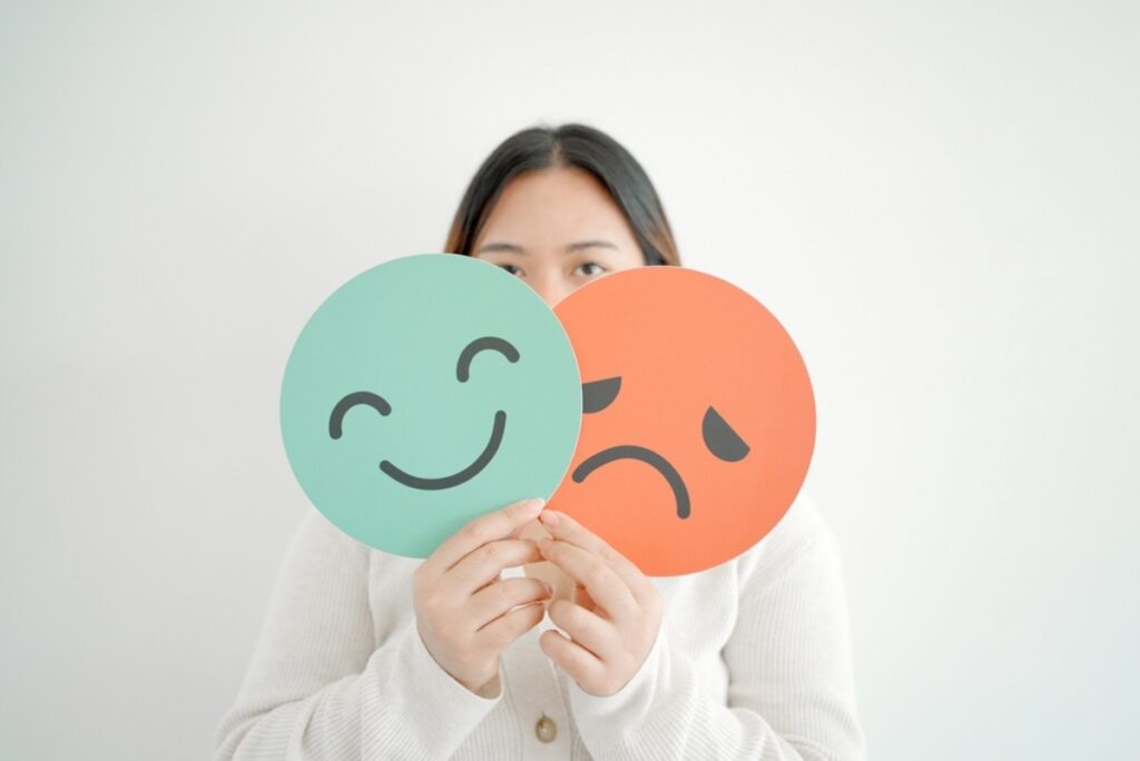 Woman holding happy and sad emoji faces illustrating the difference between an anxiety attack vs panic attack