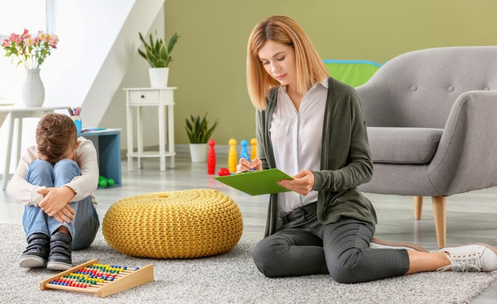 A child therapist sitting on the floor taking notes on a clipboard while a young boy sits nearby during an early autism treatment in children session