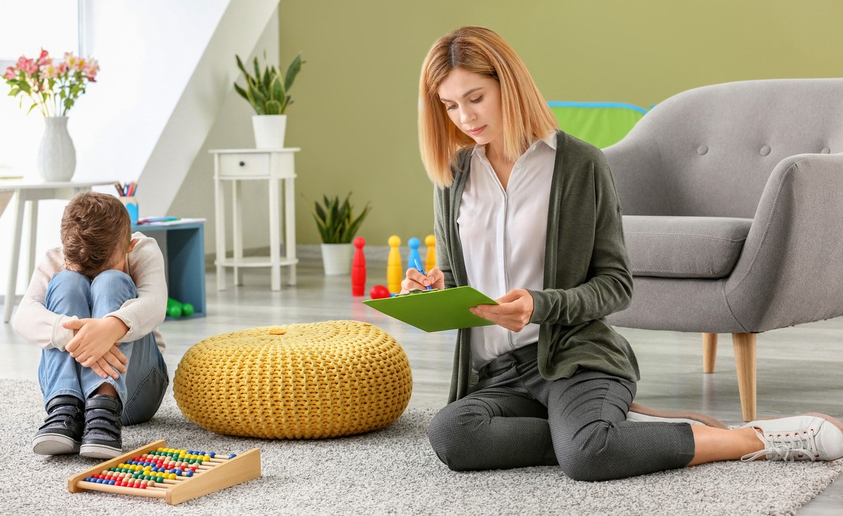 A child therapist sitting on the floor taking notes on a clipboard while a young boy sits nearby during an early autism treatment in children session